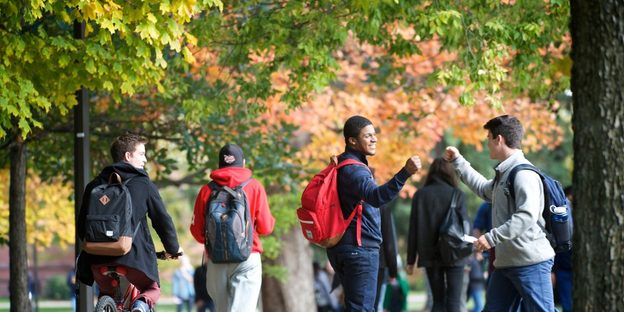 Two students greet each other as others walk along a sidewalk