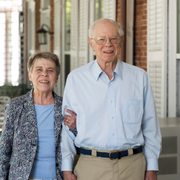 An elderly couple pose arm-in-arm, smiling